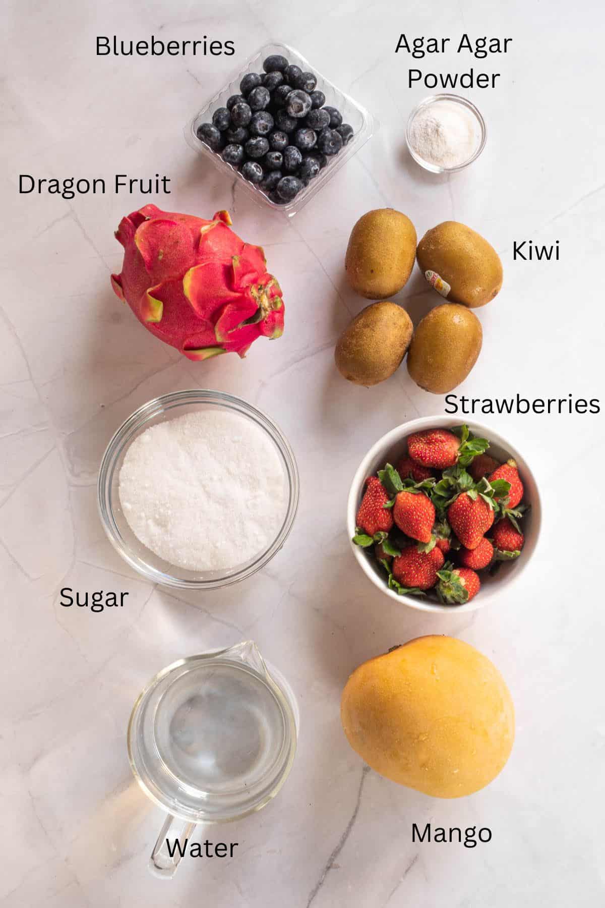 Dragon fruit, mango, strawberries, kiwis, blueberries, water, sugar and agar agar powder against a marble background.